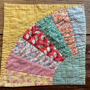 Pode incluir: Um pequeno bloco de colcha quadrado com um design vibrante em forma de leque. O bloco apresenta um fundo amarelo e um patchwork de tecidos coloridos, incluindo padrões florais e de bolinhas em vermelho, verde, laranja e rosa. O bloco de colcha é provavelmente feito à mão.