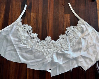May include: Ivory-colored lingerie top with delicate floral lace detailing across the neckline. The fabric appears to be a soft satin or silk, with thin straps for support. The garment is laid flat on a wooden surface.