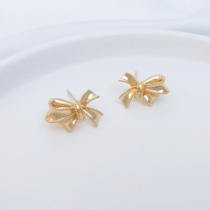 Könnte beinhalten: Ein Paar goldene Ohrstecker in Form einer Schleife. Die Schleifen sind aus strukturiertem Metall gefertigt und haben ein zartes, feminines Design.