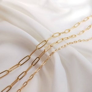 May include: Three gold chain necklaces of different lengths and thicknesses, with oval-shaped links. The chains are arranged in a staggered pattern on a white fabric background.