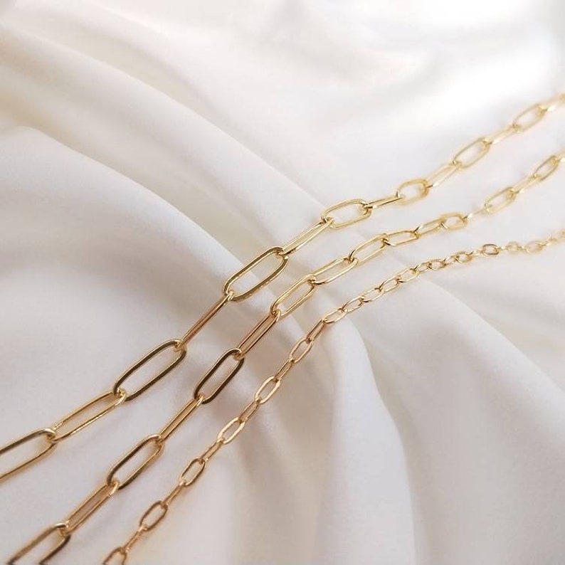 May include: Three gold chain necklaces of different lengths and thicknesses, with oval-shaped links. The chains are arranged in a staggered pattern on a white fabric background.
