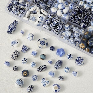 May include: A collection of blue and white ceramic beads in various shapes and sizes, including round, oval, and irregular forms. Some beads feature floral and geometric patterns. The beads are displayed in a clear plastic organizer.