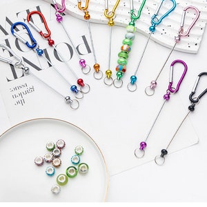 May include: Assortment of colourful metal keychains with a variety of charms, including beads, balls, and a small carabiner. The keychains are attached to a silver metal chain.