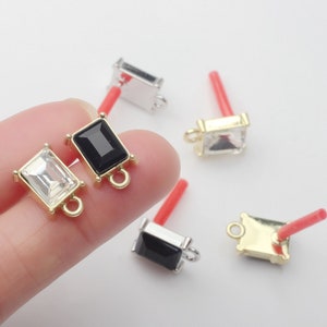May include: Close-up of several rectangular stud earrings with a post. The earrings have a gold or silver-coloured setting and a clear or black rectangular stone. The posts are red.
