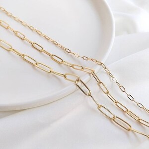 May include: Three gold chain necklaces with different sized oval links. The chains are layered on a white surface.