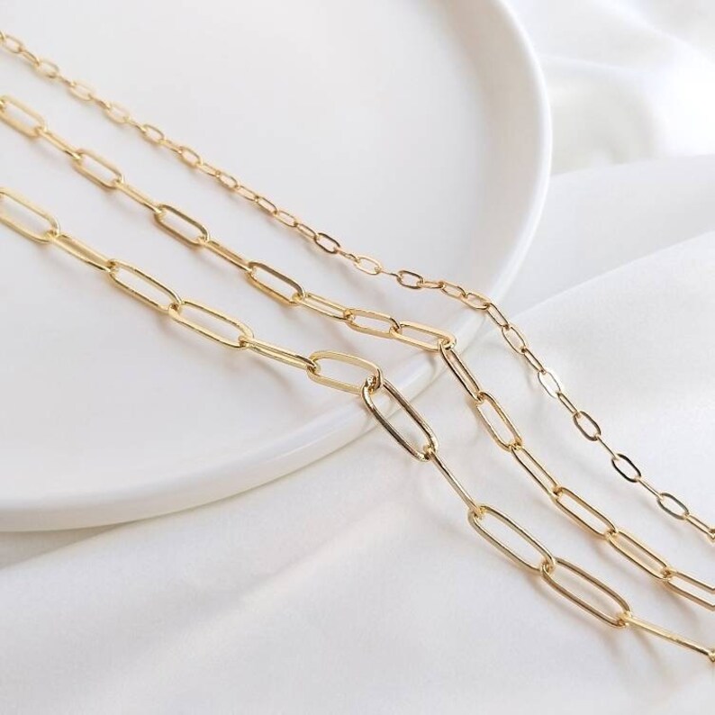 May include: Three gold chain necklaces with different sized oval links. The chains are layered on a white surface.