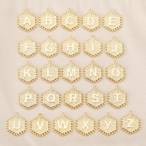 May include: Gold-colored hexagonal alphabet charms, each with a letter from A to Z. The charms have a textured border and a small loop at the top for attaching to jewelry. The background is a light beige.