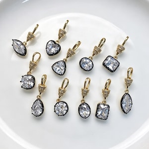 May include: A collection of ten gold-toned metal pendants featuring clear faceted crystals set in black frames. Each pendant includes a gold-toned bail with small embedded stones, showcasing various shapes including heart, pear, oval, and rectangular.