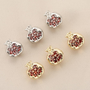 Può includere: Sei ciondoli a forma di melograno. Tre sono argentati e tre dorati. Ogni ciondolo è riempito di piccole pietre preziose rotonde rosse. I ciondoli sono disposti su una superficie beige chiaro.