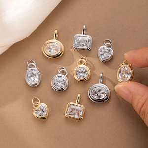 Puede incluir: Una colección de pequeños dijes de joyería con piedras facetadas transparentes engastadas en marcos de metal en tonos oro y plata. Los dijes presentan varias formas, incluyendo redondas, ovaladas, rectangulares y de corazón, con argollas.