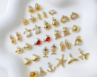 May include: A collection of gold-colored charms in various marine life shapes, including starfish, seahorses, fish, and shells. Some charms feature clear or red gemstones. The charms are arranged on a white, slightly textured surface.