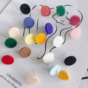 Pode incluir: Conjunto de 17 cabochões redondos, planos e coloridos de resina com um furo no centro. Os cabochões são de várias cores, incluindo vermelho, rosa, azul, verde, amarelo, laranja, castanho, preto e branco. Os cabochões estão dispostos numa superfície branca.