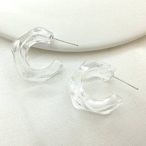 May include: A pair of clear, transparent hoop earrings. The earrings have an unusual, irregular shape and a silver post. The earrings are displayed on a white surface.