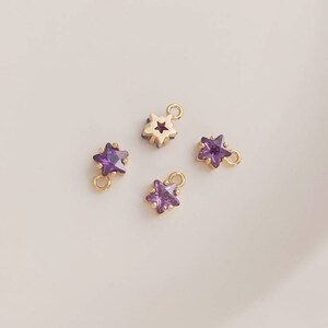 May include: Four gold star-shaped charms with purple crystal accents. Two charms have a loop at the top for hanging, and two charms have a loop on the side.
