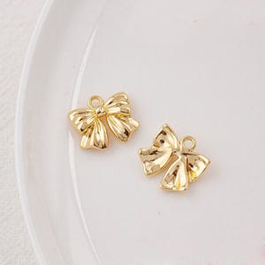 May include: Two gold-coloured metal bow charms. The bows are detailed with a textured surface and have a loop at the top for attaching to jewellery or other crafts.