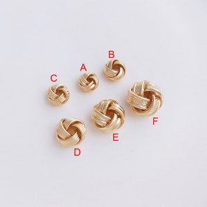 Op de afbeelding: Zes gouden metalen knoop kralen in verschillende maten. De kralen zijn in een halve cirkel op een wit oppervlak gerangschikt.