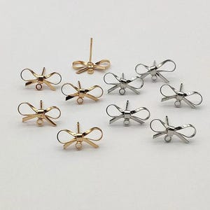 Pode incluir: Conjunto de peças para brincos em forma de laço, em ouro e prata. As peças são de metal e têm um pequeno laço na parte superior para prender a um poste de brinco.