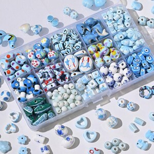 Puede incluir: Un organizador de plástico transparente lleno de una variedad de cuentas de cerámica azules y blancas. Las cuentas tienen varias formas, incluyendo redondas, cuadradas y florales, con patrones decorativos. Algunas cuentas tienen caras sonrientes o formas de delfines.