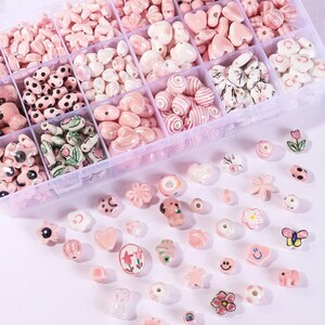 Peut inclure: Un organisateur en plastique transparent rempli de diverses perles de céramique roses. Les perles ont différentes formes, notamment des cœurs, des fleurs et des étoiles. Certaines perles ont des détails peints, tels que des rayures et des points.