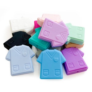 Op de afbeelding: Een verzameling kleurrijke siliconen kralen in de vorm van medische scrubs. De kralen zijn in verschillende pastelkleuren, waaronder blauw, roze, paars en groen. Elke kraal heeft een hart en zakdetails. Ideaal voor knutselen.