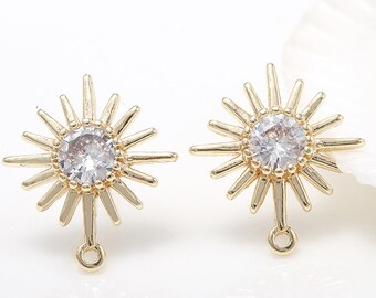 May include: A pair of gold earrings with a sunburst design and a clear crystal in the center. The earrings have a drop design with a small loop at the bottom.