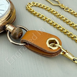 May include: Close-up of a gold pocket watch with a white face and black Roman numerals. A tan leather strap with a gold clasp and chain is attached. The watch and chain are on a light gray surface.