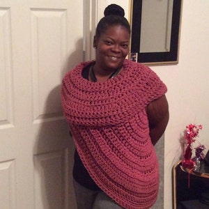 May include: A woman wearing a pink crocheted shawl with a cowl neck. The shawl is draped over her shoulders and is made with a simple stitch pattern.