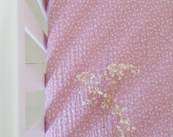 May include: A pink cot sheet with a white floral pattern. The sheet is made of soft fabric and is partially visible in a cot. A sprig of dried white flowers rests on the sheet.