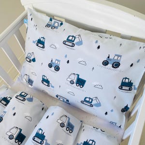 Peut inclure: Ensemble de literie pour bébé blanc, orné d'un motif de véhicules de chantier bleus, de nuages et d'arbres. L'ensemble comprend un oreiller et d'autres pièces, toutes avec le même motif répétitif. Les véhicules comprennent des camions, des tracteurs et des excavatrices.