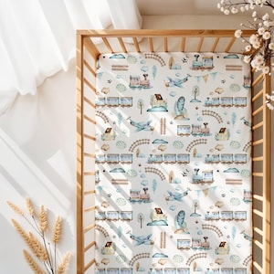 May include: A white fitted cot sheet featuring a repeating pattern of trains, aeroplanes, houses, trees, and clouds in blue, brown, and green. The sheet is inside a light-coloured wooden cot.