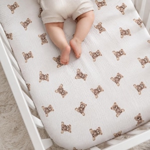 Peut inclure: Un drap de lit blanc avec un motif répétitif de petits oursons bruns. Le tissu présente un aspect texturé et froissé. Une paire de pieds nus et le bas des jambes portant un short côtelé beige clair reposent sur le drap.