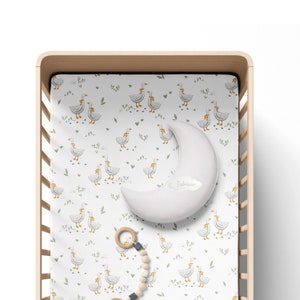 May include: A white cot sheet with a pattern of geese and leaves. A white crescent moon shaped pillow with the word "Fabricee" printed on it is on the sheet. A wooden toy with a ring and beads is on the sheet.