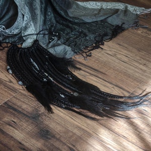 Könnte beinhalten: Schwarze Dreadlocks mit silbernen Perlen, die von einem grau-weißen gemusterten Schal auf einem Holzboden hängen.