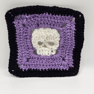 May include: A crocheted square with a purple centre and a black border. A white skull is crocheted in the centre of the square.