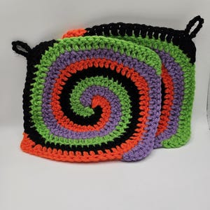 May include: Two square, handmade crochet potholders. Each potholder features a spiral design with orange, black, green, and purple yarn. The potholders have a loop for hanging. The overall theme is Halloween.