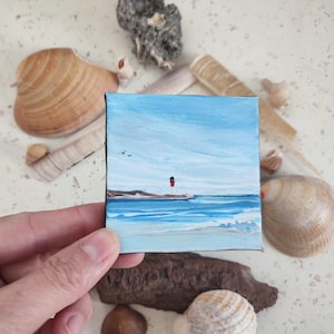 Può includere: Un piccolo dipinto su tela che rappresenta un paesaggio marino con un faro rosso e bianco su una piccola isola in lontananza. Il cielo è blu con nuvole bianche e l'acqua è azzurra con onde bianche.