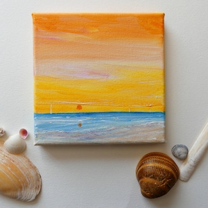 Peut inclure: Une petite peinture sur toile représentant un coucher de soleil sur une mer bleue. Le ciel est un dégradé d'orange, de jaune et de rose. La mer est d'un bleu clair avec des vagues blanches. La peinture est encadrée d'une bordure blanche.
