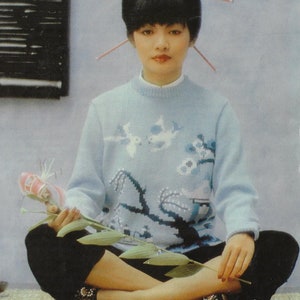 May include: A woman wearing a light blue jumper with a floral design and black trousers. She is sitting with her legs crossed and holding a pink flower.