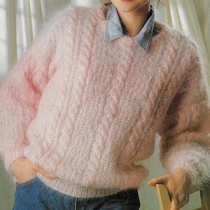 Puede incluir: Un suéter rosa claro y difuso con cuello redondo y detalles de punto trenzado. El suéter tiene mangas largas y se usa sobre una camisa de mezclilla. La textura suave y el color claro le dan al suéter una apariencia delicada.