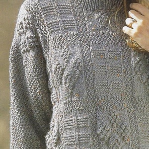 Könnte beinhalten: Ein grauer Strickpullover mit einem strukturierten Muster. Der Pullover hat einen Rundhalsausschnitt und lange Ärmel.