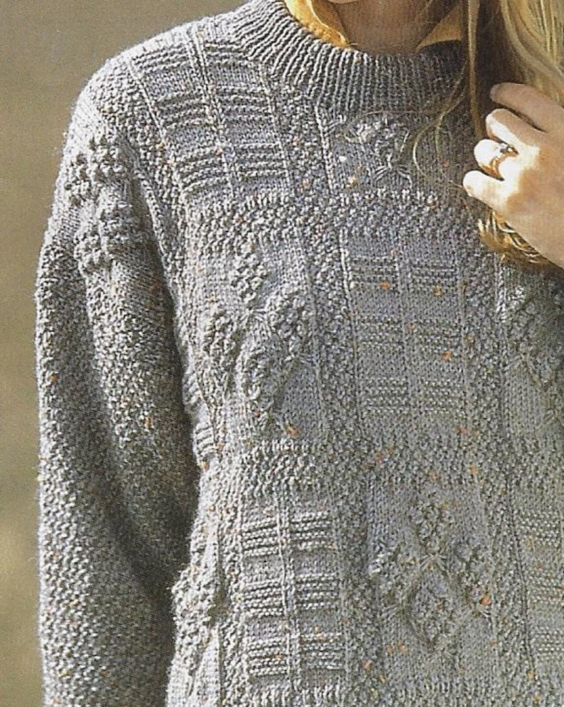 K&ouml;nnte beinhalten: Ein grauer Strickpullover mit einem strukturierten Muster. Der Pullover hat einen Rundhalsausschnitt und lange &Auml;rmel.
