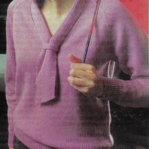 May include: A pink knitted sweater with a V-neck and a bow tie detail.