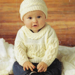 May include: A baby wearing a white knitted sweater and hat with a pom-pom on top. The baby is sitting on a wooden floor with a white blanket behind them.