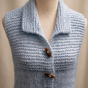 May include: A light blue knitted vest with a collared neckline and wooden toggle buttons. The vest is displayed on a mannequin and features a textured knit pattern. The buttons are a warm brown color, contrasting with the cool blue of the vest.