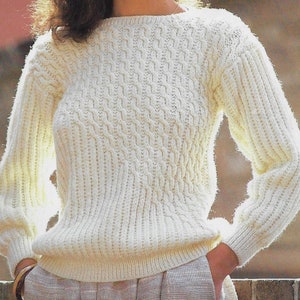 Puede incluir: Un suéter de punto blanco con un patrón de trenza. El suéter tiene un cuello redondo y mangas largas.
