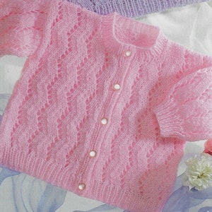 以下が含まれることがあります： 繊細なレース模様とボタン留めが施されたピンクのベビー用ニットカーディガンです。