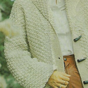 May include: A cream-colored knitted cardigan with a button closure and a textured pattern. The cardigan is worn over a white shirt and brown pants.
