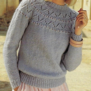 Puede incluir: Un suéter de punto azul claro con un delicado patrón de encaje alrededor del cuello y los puños. El suéter tiene un corte holgado y mangas largas.