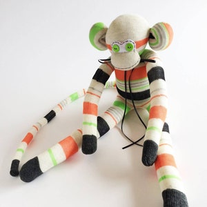 May include: A handmade sock monkey toy with a white, orange, green, and black striped body. The monkey has a long tail and black eyes with green pupils.