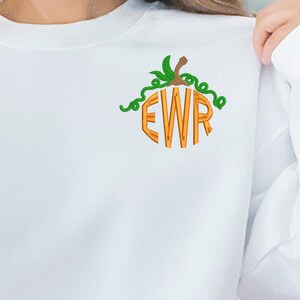 Pode incluir: Camisola branca com um monograma em forma de abóbora das letras "EWR" em laranja com videiras verdes.